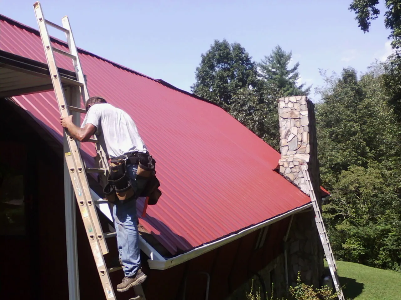 Roofer on a ladder beside a red metal roof and stone chimney — Soffit & Fascia Repair by Roofing Brothers in Long Grove